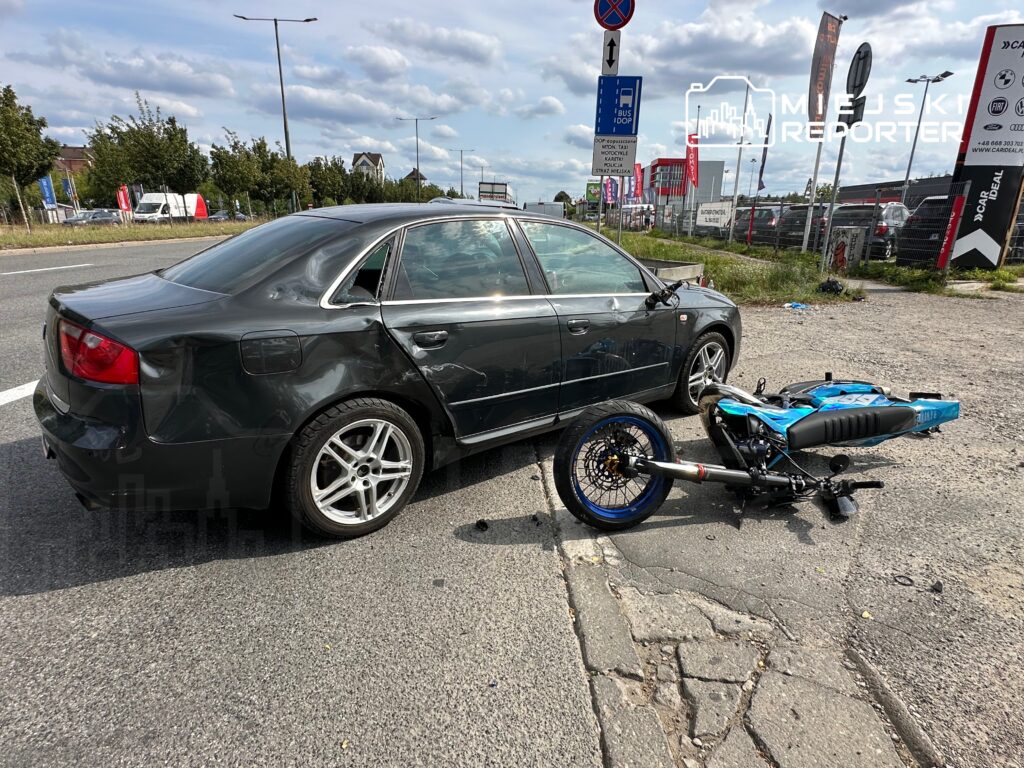 Czarny samochód osobowy z uszkodzeniami na karoserii stoi obok przewróconego motocykla w miejskim otoczeniu.