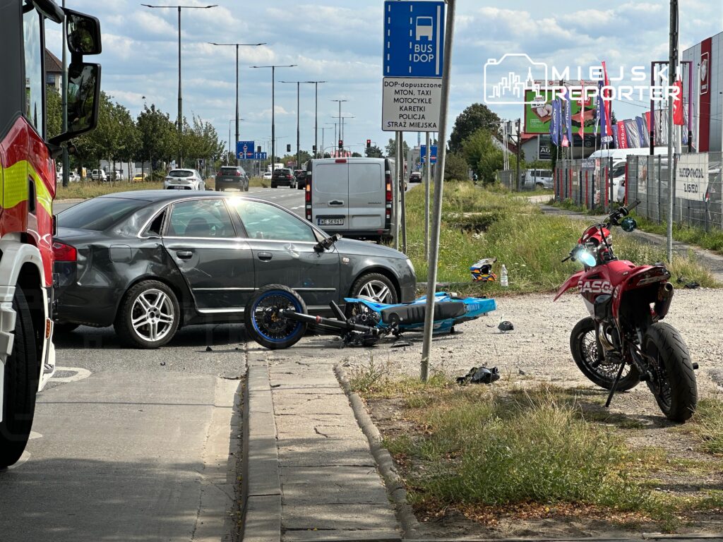 Zderzenie samochodu osobowego z motocyklem na drodze, obok stojących pojazdów i znaków drogowych.