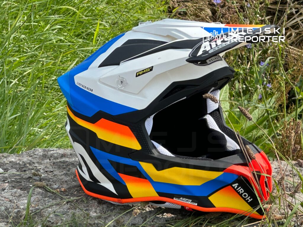 Kask motocyklowy w kolorach niebieskim, pomarańczowym i czarnym leży na kamieniu, otoczony zieloną trawą.