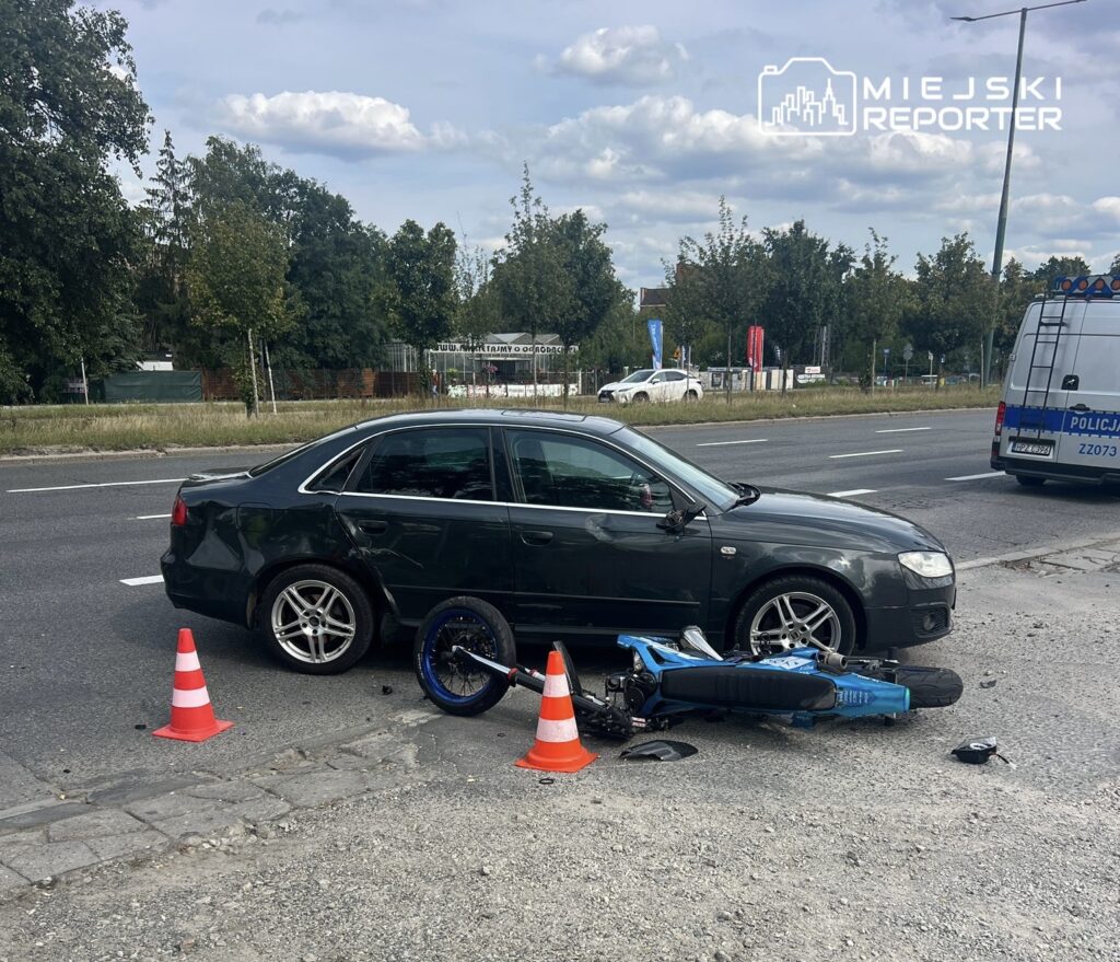 Czarny samochód osobowy stoi na poboczu drogi obok przewróconego niebieskiego motocykla, otoczonego pachołkami.