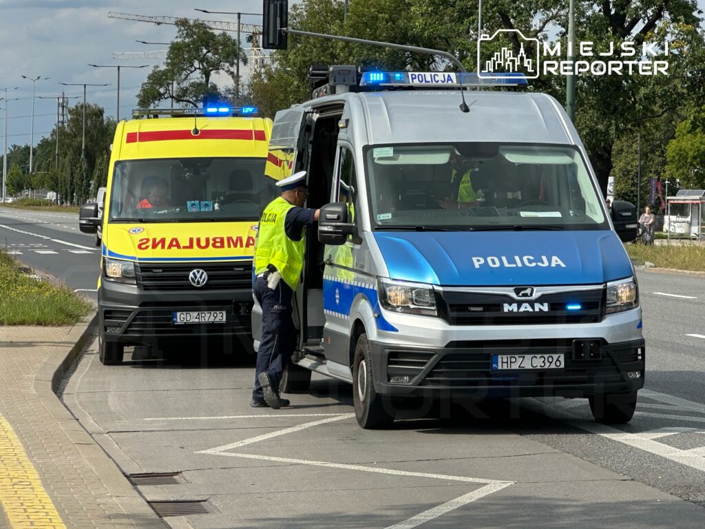 Policjant rozmawia z kierowcą ambulansu na miejskiej ulicy, obok stoi radiowóz z włączonymi sygnałami świetlnymi.
