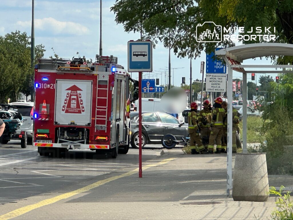 W tle widoczny wóz strażacki z włączonymi sygnałami świetlnymi, a w pierwszym planie strażacy w mundurach omawiają sytuację przy uszkodzonym pojeździe.