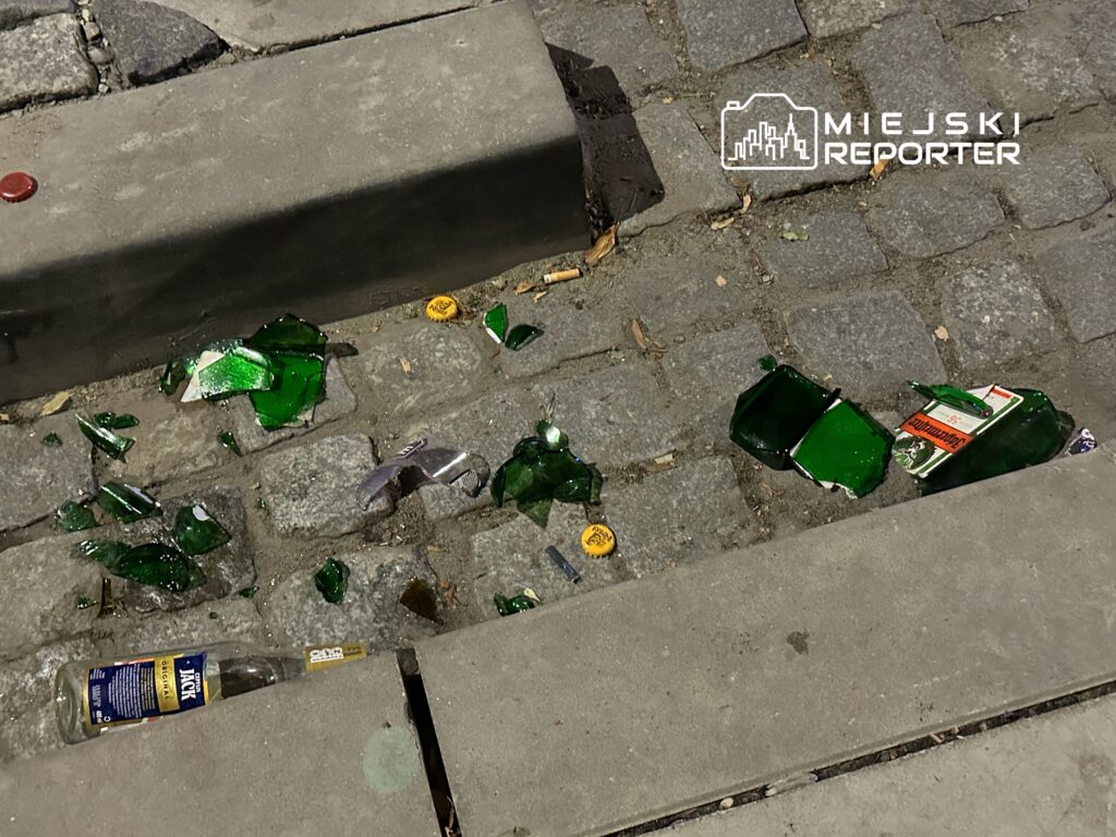 Szkło z rozbitych butelek leży na chodniku, wśród kamieni brukowych, obok kapsli i resztek etykiet.