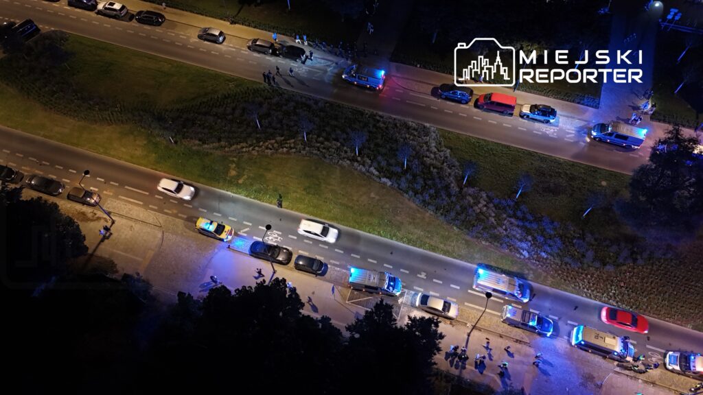 Grupa policjantów interweniuje na oświetlonej ulicy, obok zaparkowanych samochodów, z włączonymi sygnałami świetlnymi.