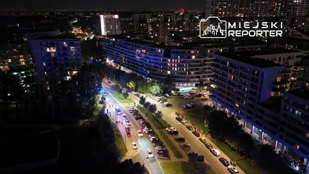Nocna panorama miasta z widokiem na oświetloną ulicę, gdzie widać radiowozy i grupę ludzi.