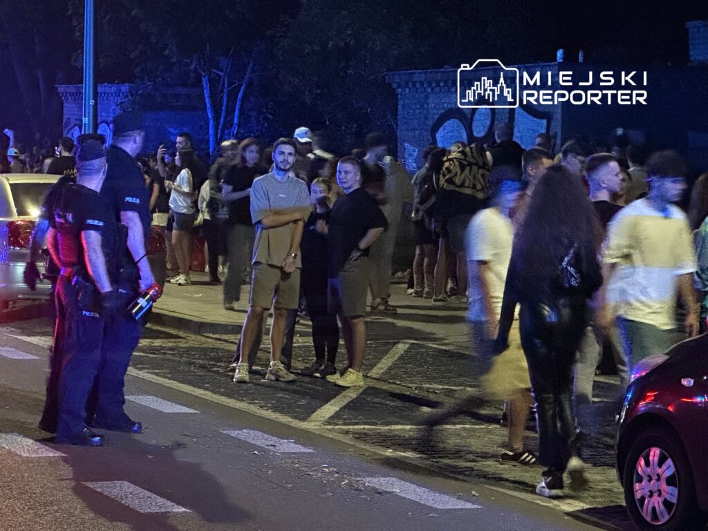 Grupa ludzi gromadzi się na ulicy, a policjanci w mundurach monitorują sytuację w tle.