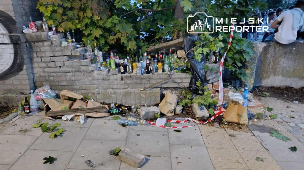 Zniszczony mur z wystającymi butelkami po alkoholu, porozrzucane odpady i liście na chodniku.