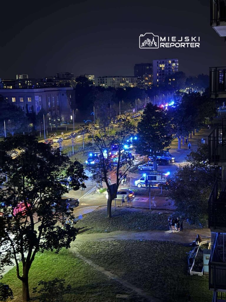 Na ulicy w nocy, pojazdy policyjne z włączonymi sygnałami świetlnymi gromadzą się wokół zdarzenia, a przechodnie obserwują sytuację.