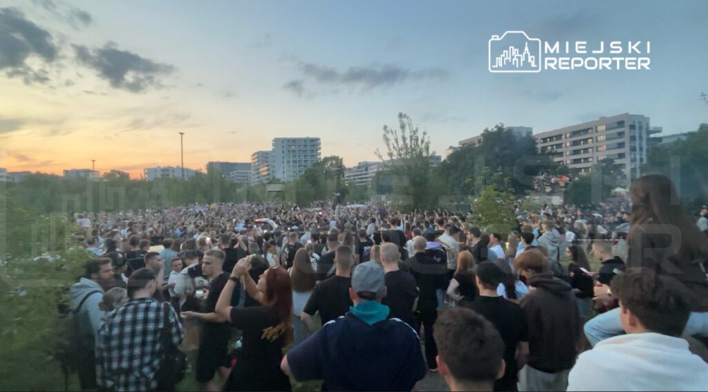 Tłum ludzi zgromadzony na świeżym powietrzu, w otoczeniu drzew i nowoczesnych budynków, podczas zachodu słońca.