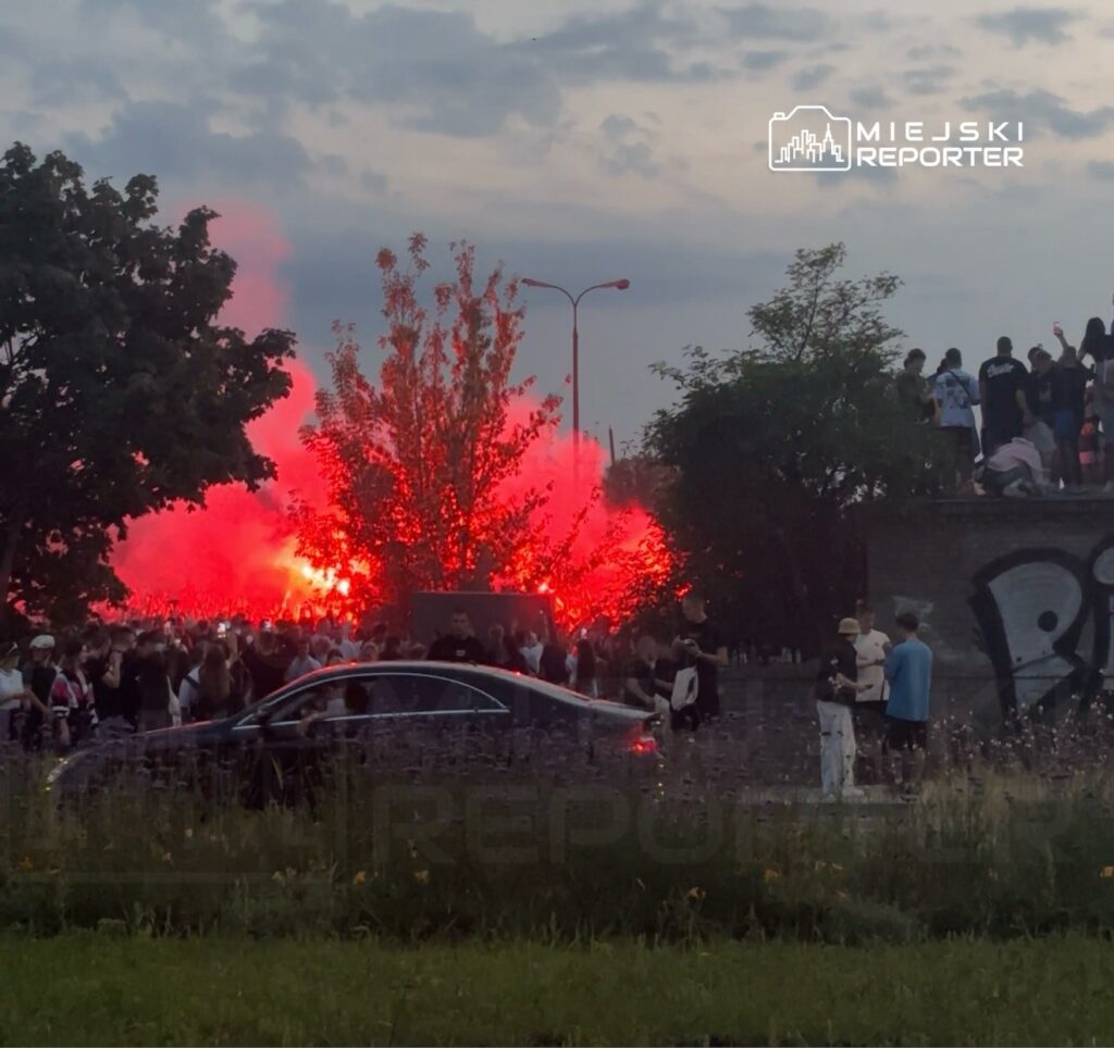 Tłum ludzi gromadzi się w otoczeniu ognia i dymu, na tle drzew i graffiti, w atmosferze intensywnego świętowania.
