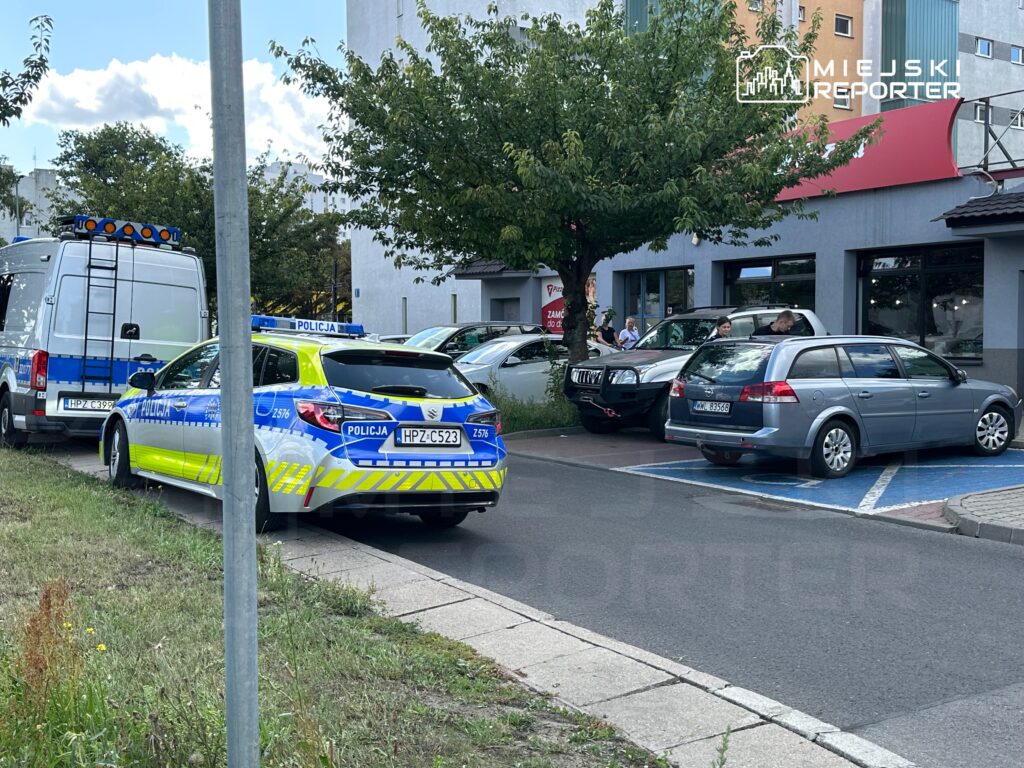 Radiowóz policyjny i furgonetka z niebieskimi światłami stoją na parkingu obok budynku, obok kilku zaparkowanych samochodów.