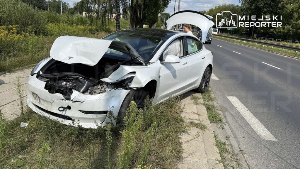 Uszkodzony biały samochód osobowy z otwartą maską stoi na poboczu drogi, obok niego kobieta sprawdzająca stan pojazdu.