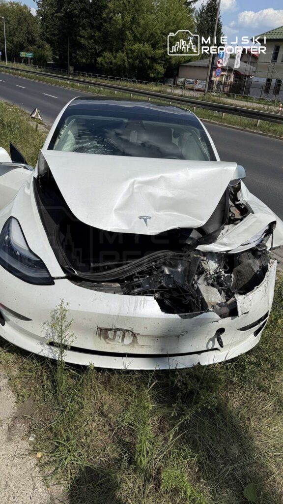 Uszkodzony biały samochód osobowy marki Tesla z wgniecioną maską stoi na poboczu drogi, otoczony wysoką trawą.