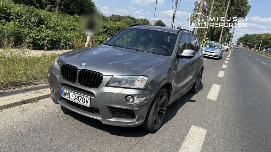 Uszkodzony samochód osobowy marki BMW zaparkowany na poboczu drogi, obok policyjny radiowóz. W tle widoczna zieleń.