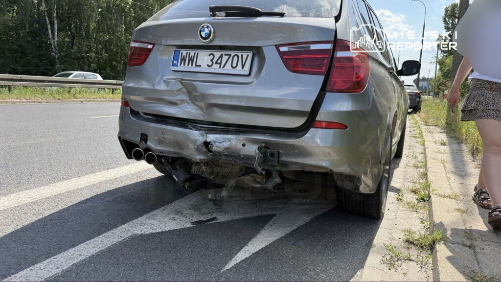 Uszkodzony samochód marki BMW z wgniecionym tyłem stoi na poboczu drogi, obok przechodząca kobieta.