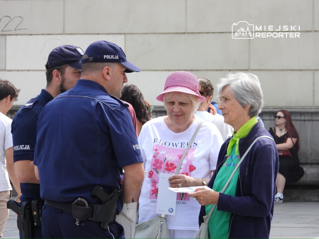 Dwaj policjanci rozmawiają z dwiema starszymi kobietami na ulicy, w tle widoczni przechodnie.