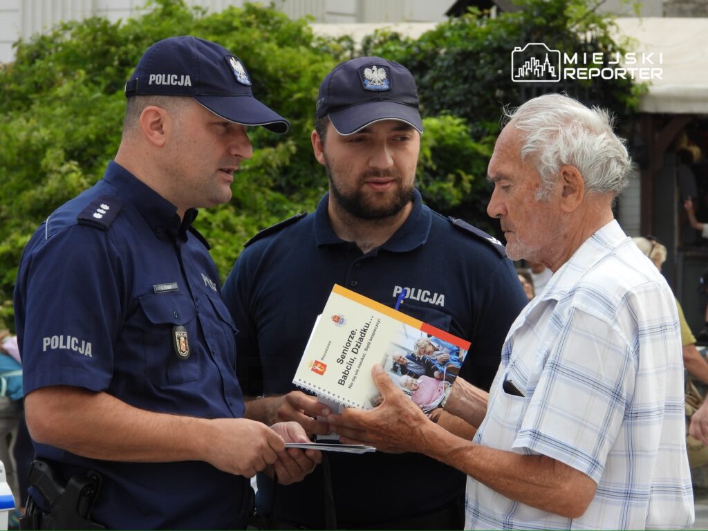 Dwaj policjanci rozmawiają z starszym mężczyzną, trzymającym broszurę, na tle zieleni i tłumu.