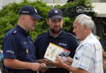 Dwaj policjanci rozmawiają z starszym mężczyzną, trzymającym broszurę, na tle zieleni i tłumu.