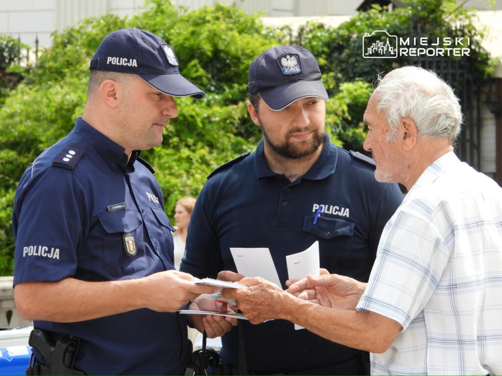 Dwaj policjanci rozmawiają z starszym mężczyzną, trzymając w rękach dokumenty, w zielonym otoczeniu.