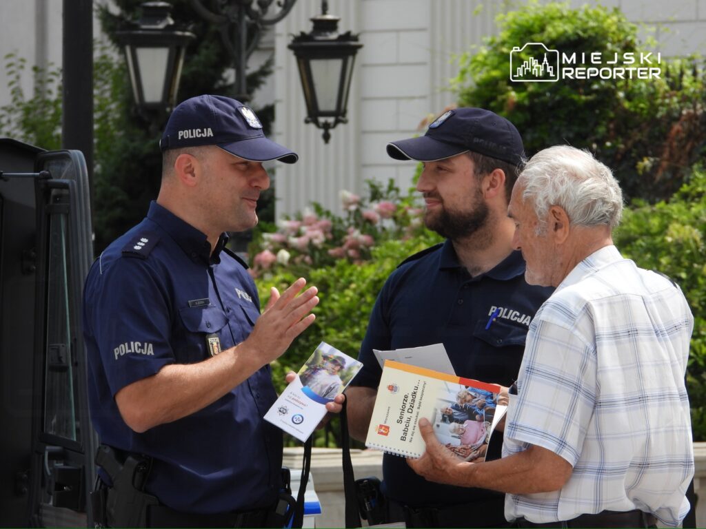 Dwaj policjanci rozmawiają z starszym mężczyzną, trzymając ulotki, w tle kwitnące krzewy.