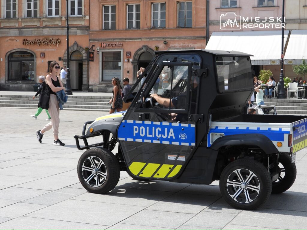 Kobieta w czarnym topie i jasnych spodniach spaceruje obok policyjnego pojazdu na placu w mieście.
