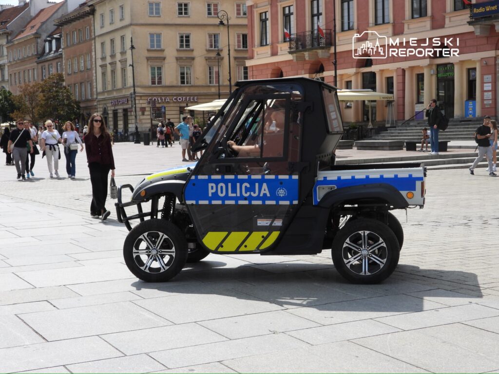 Pojazd policyjny na miejskim placu, obok przechodzących ludzi w letnich ubraniach.