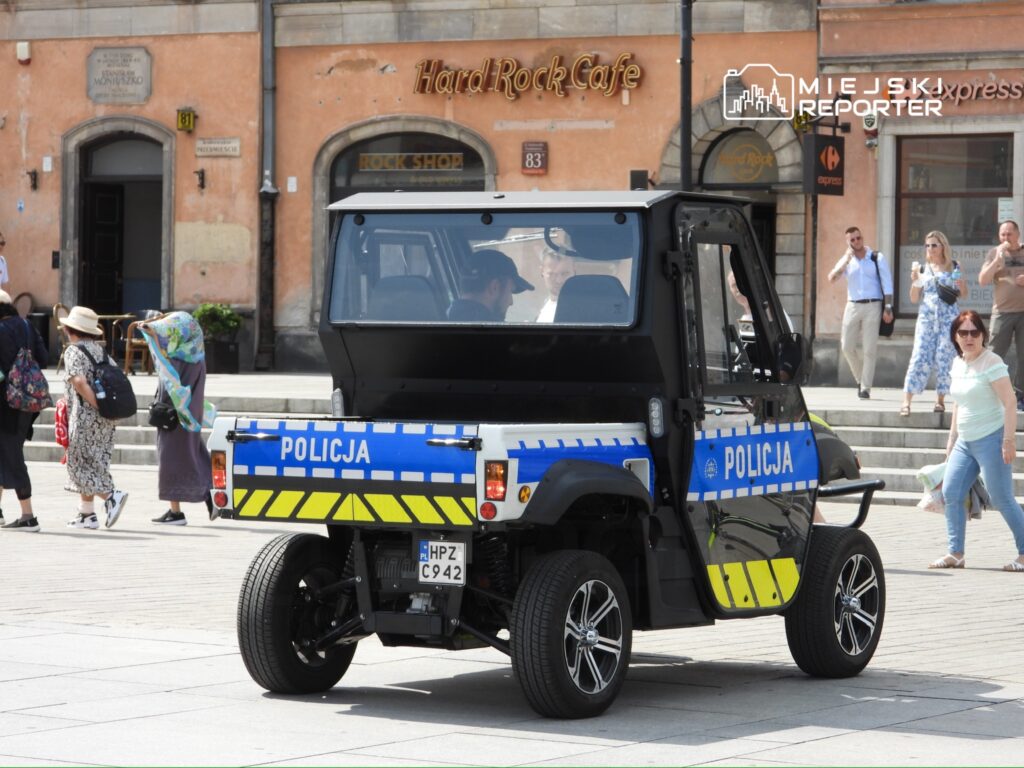 Policyjny pojazd patrolowy z napisem "POLICJA" porusza się po ruchliwym placu, obok przechodniów i lokali.