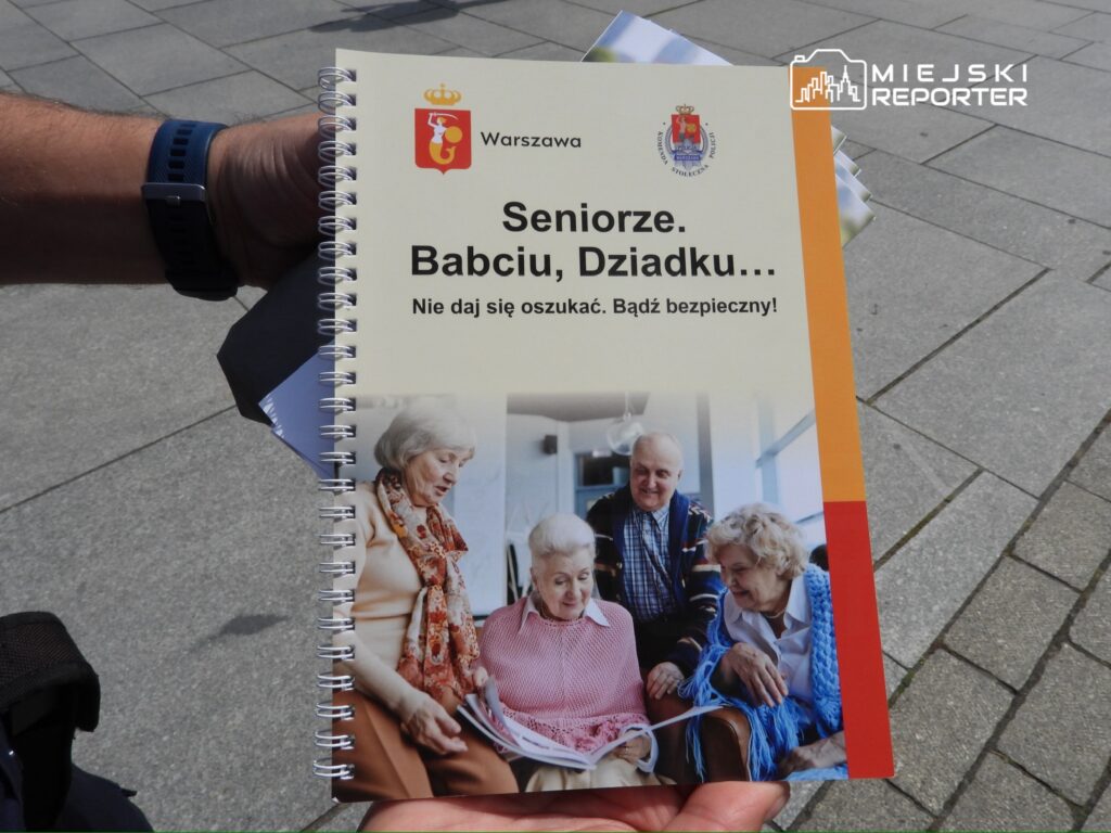 Osoba trzymająca broszurę z napisem "Seniorze. Babciu, Dziadku... Nie daj się oszukać. Bądź bezpieczny!" na tle placu.
