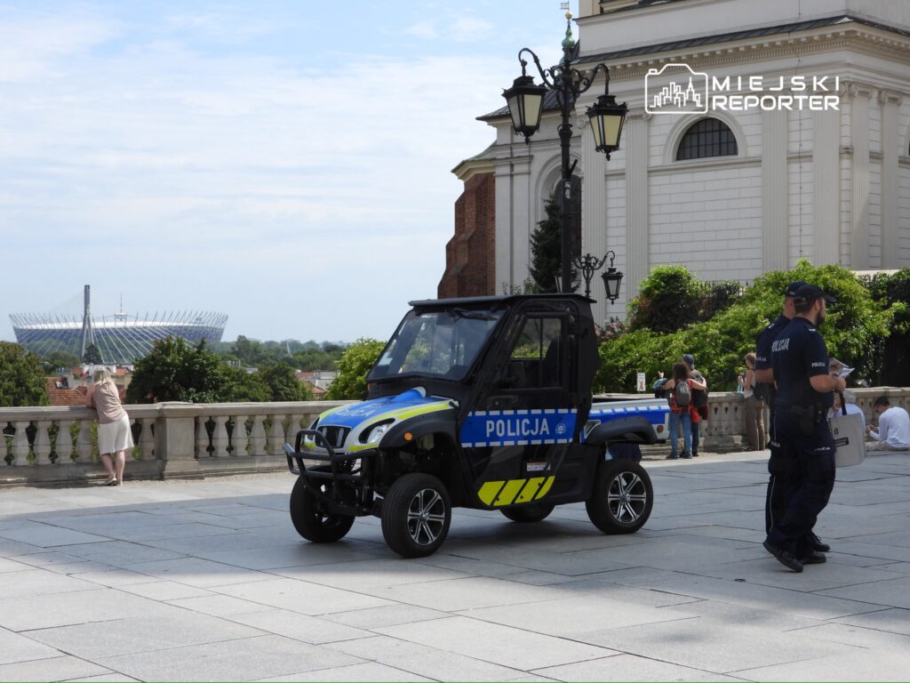 Funkcjonariusze policji rozmawiają obok policyjnego pojazdu w parku, z widokiem na stadion w tle.