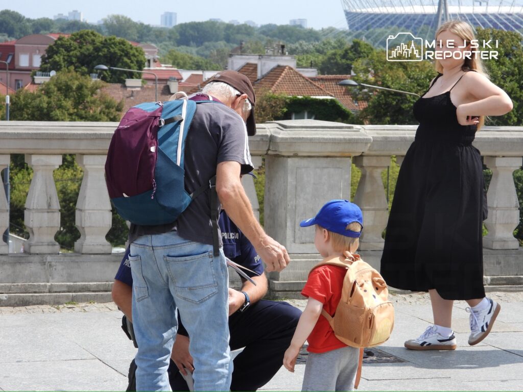 Mężczyzna w czapce rozmawia z dzieckiem w niebieskiej czapce, podczas gdy kobieta w czarnej sukience stoi obok.