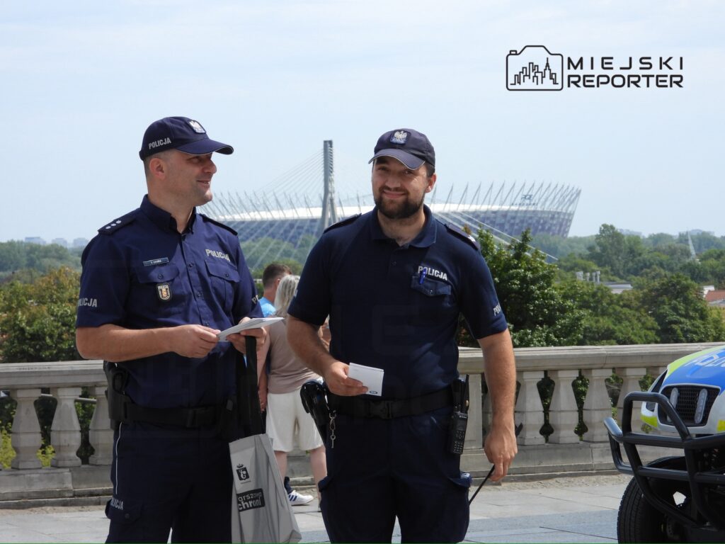 Dwóch policjantów rozmawia na tle stadionu, trzymając notatki w rękach. W tle widoczni przechodnie i zieleń.