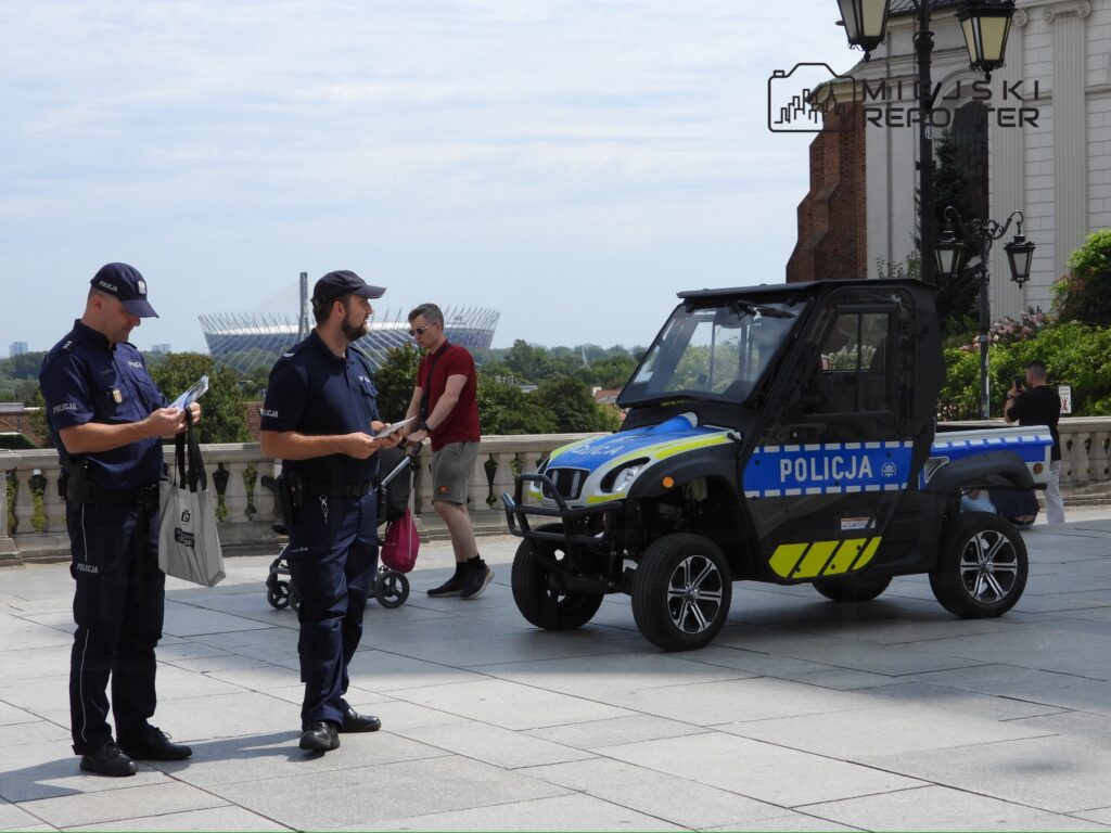 Dwóch policjantów przegląda dokumenty obok radiowozu na placu, w tle stadion i zieleń.
