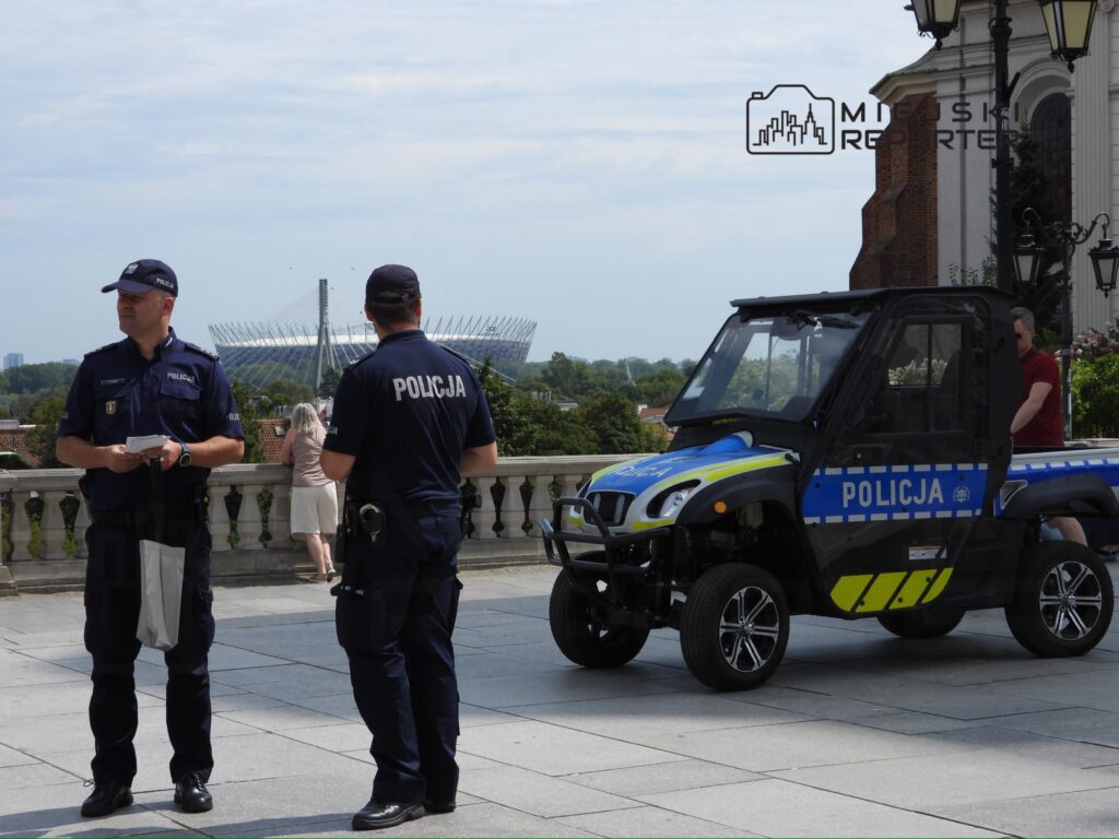 Dwaj policjanci w mundurach stoją na tarasie, obok pojazdu policyjnego, w tle widoczny stadion.
