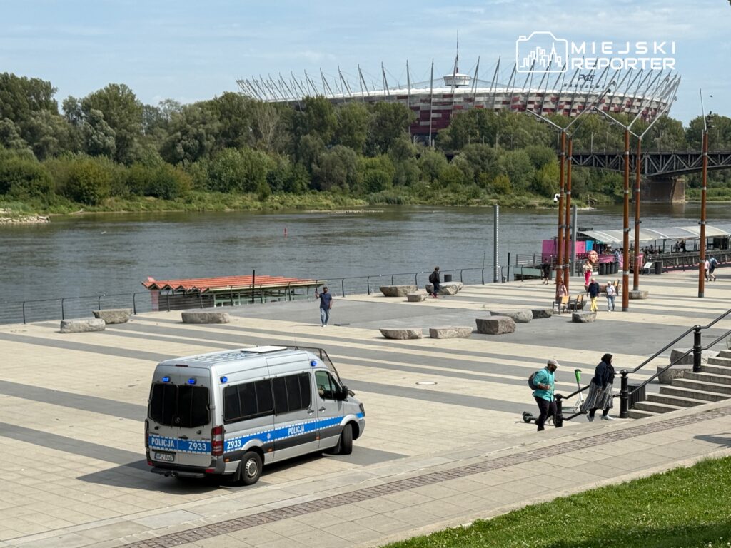 Policyjny samochód zaparkowany na bulwarze nad rzeką, w tle stadion z charakterystycznym dachem.