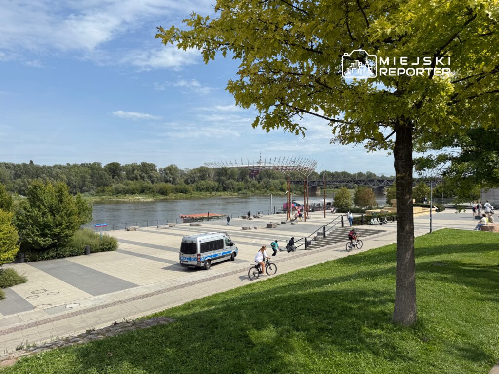 Rowerzyści poruszają się wzdłuż bulwaru nad rzeką, obok parku z drzewami i nowoczesnych elementów architektury.