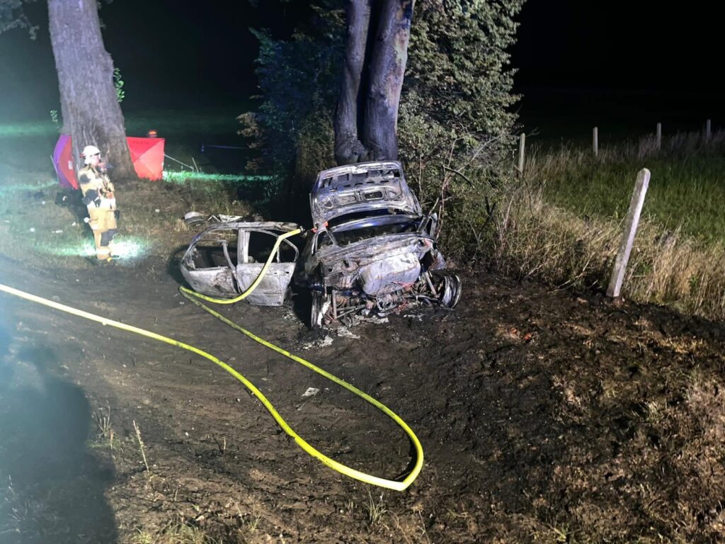 Zniszczony samochód po wypadku na poboczu drogi, obok stoi strażak z wężem gaśniczym.
