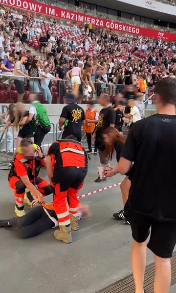 Ratownicy medyczni udzielają pomocy osobie leżącej na ziemi na tle tłumu ludzi w stadionie.