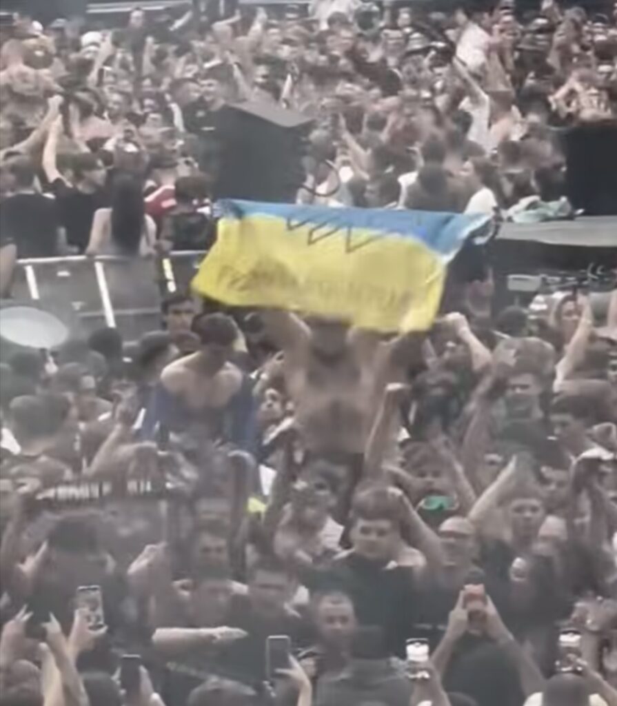 Tłum ludzi na koncercie, mężczyzna trzymający flagę Ukrainy, wszyscy wiwatują i robią zdjęcia.
