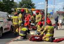 Zespół ratowników medycznych i strażaków udziela pomocy poszkodowanemu na chodniku obok zaparkowanej karetki.