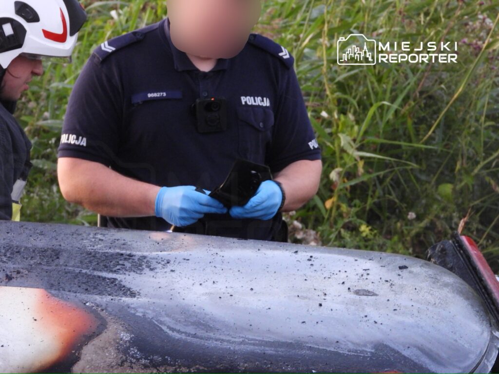 Policjant w niebieskim mundurze przeszukuje przedmioty na uszkodzonym pożarem samochodzie w zaroślach.