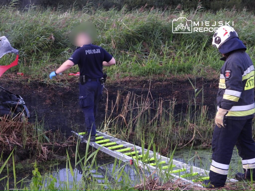 Policjant w rękawiczkach bada miejsce wypadku obok wraku samochodu, a strażak obserwuje sytuację z boku.
