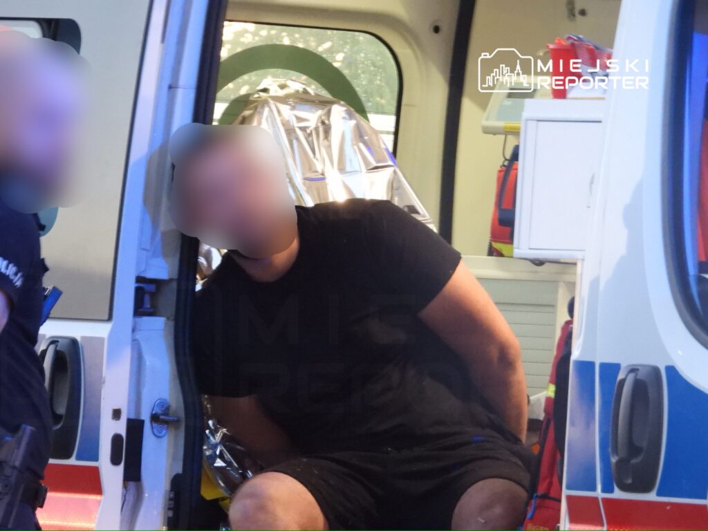 Mężczyzna w czarnej koszulce siedzący w karetce, owinięty w srebrną folię ratunkową, przy asyście ratowników.