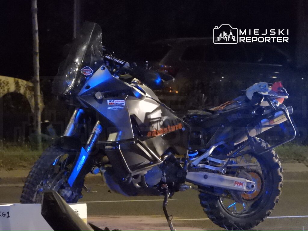 Motocykl terenowy z dużą szybą i oświetleniem, zaparkowany na drodze w nocy. W tle widoczne samochody.