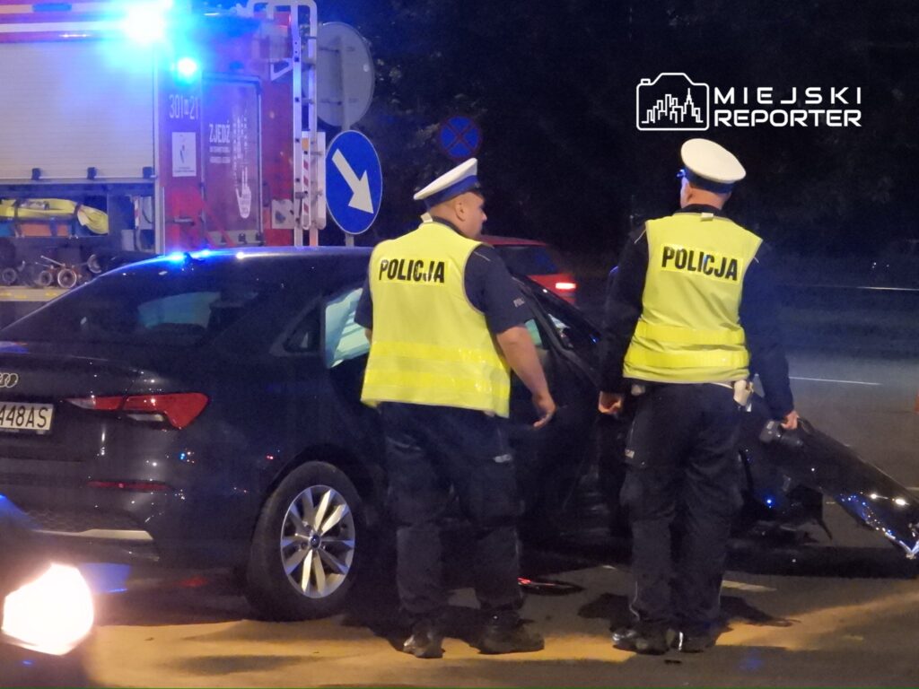 Dwaj policjanci w odblaskowych kamizelkach rozmawiają obok uszkodzonego samochodu w nocy, w tle widać woz strażacki.