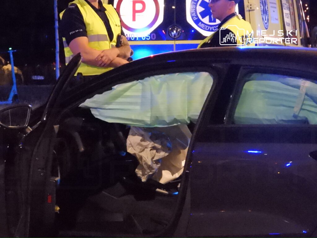 Policjant w kamizelce odblaskowej stoi obok samochodu z otwartymi drzwiami, w którym widać poduszki powietrzne.