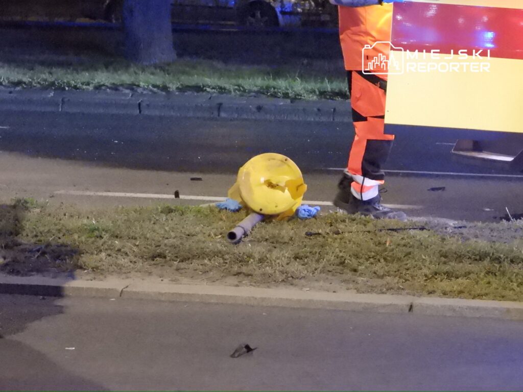 Pracownik w pomarańczowym uniformie stoi obok uszkodzonego znaku drogowym na trawniku przy jezdni.