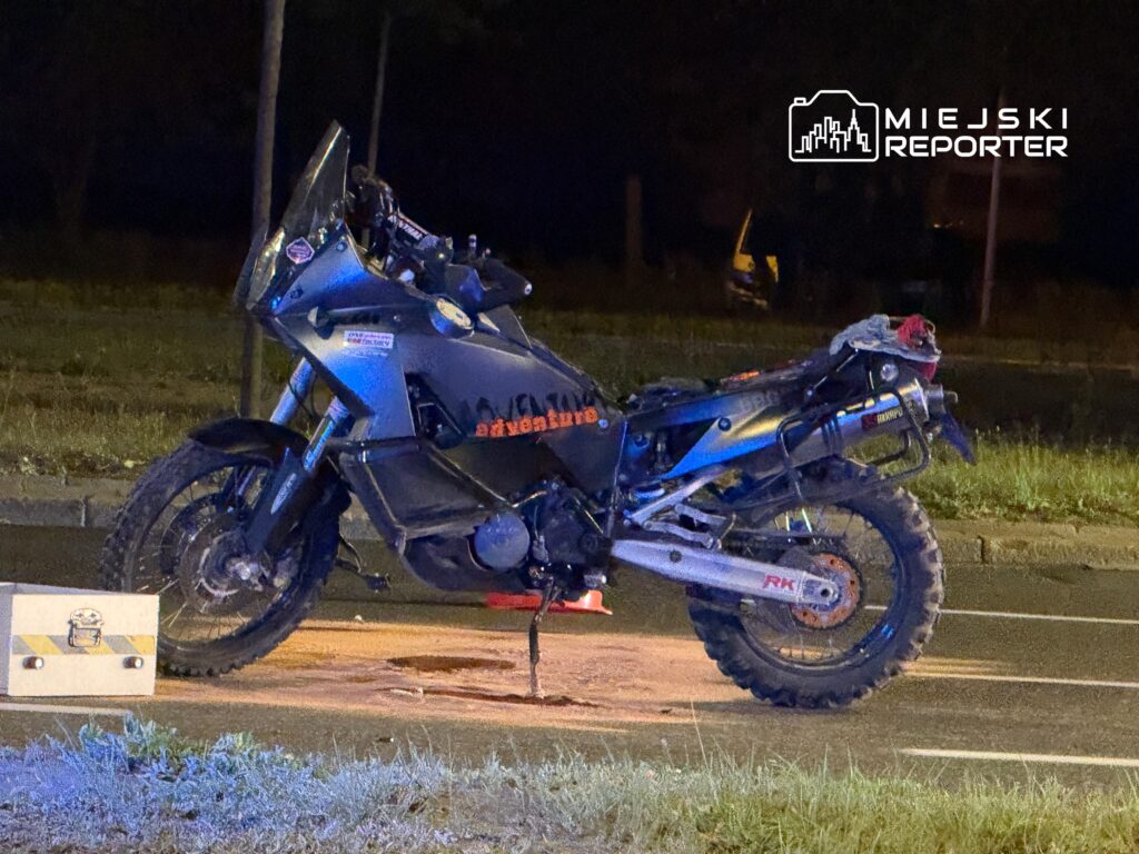 Motocykl terenowy zaparkowany na poboczu drogi, obok metalowa skrzynka. Nocne oświetlenie.