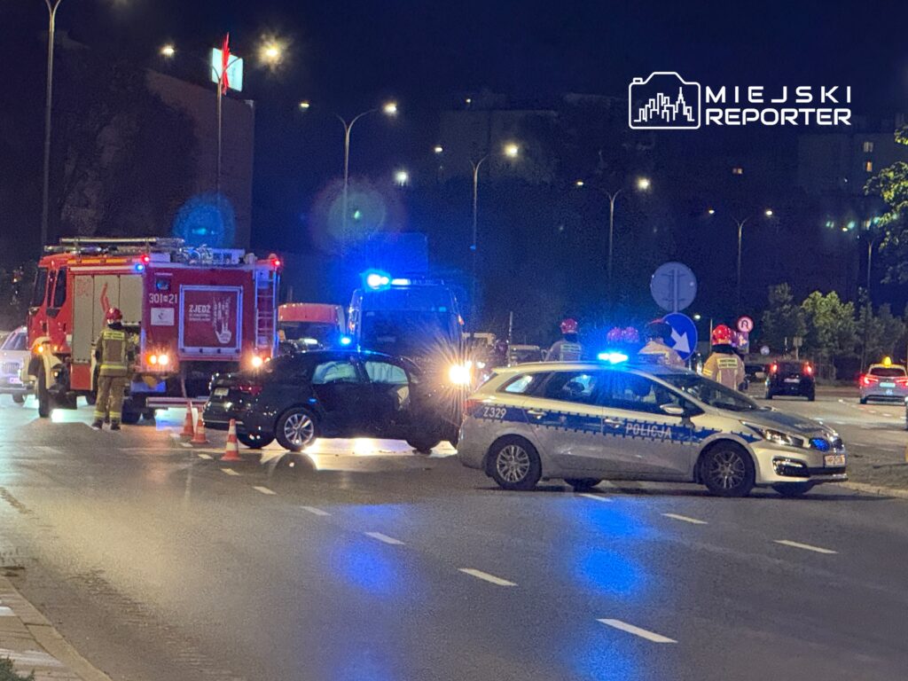 Na skrzyżowaniu nocą strażacy i policjanci zabezpieczają miejsce zdarzenia, wokół ustawione pachołki.