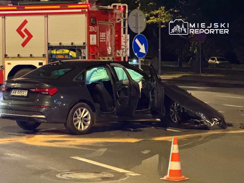 Czarny samochód osobowy z uszkodzoną karoserią stoi na skrzyżowaniu, obok wozu strażackiego.