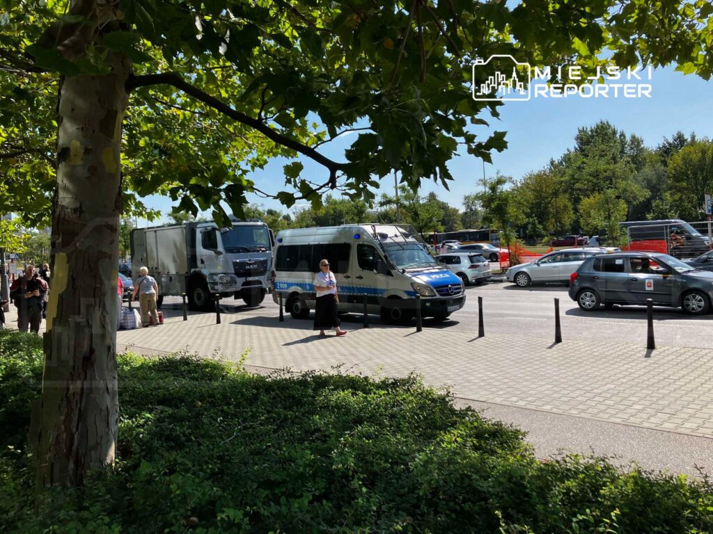 Grupa osób stoi na chodniku obok zaparkowanych pojazdów, w tym furgonetki policyjnej i ciężarówki, w słoneczny dzień.